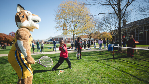 A child plays tennis with Boomer the Bobcat mascot on the Quinnipiac quad grass